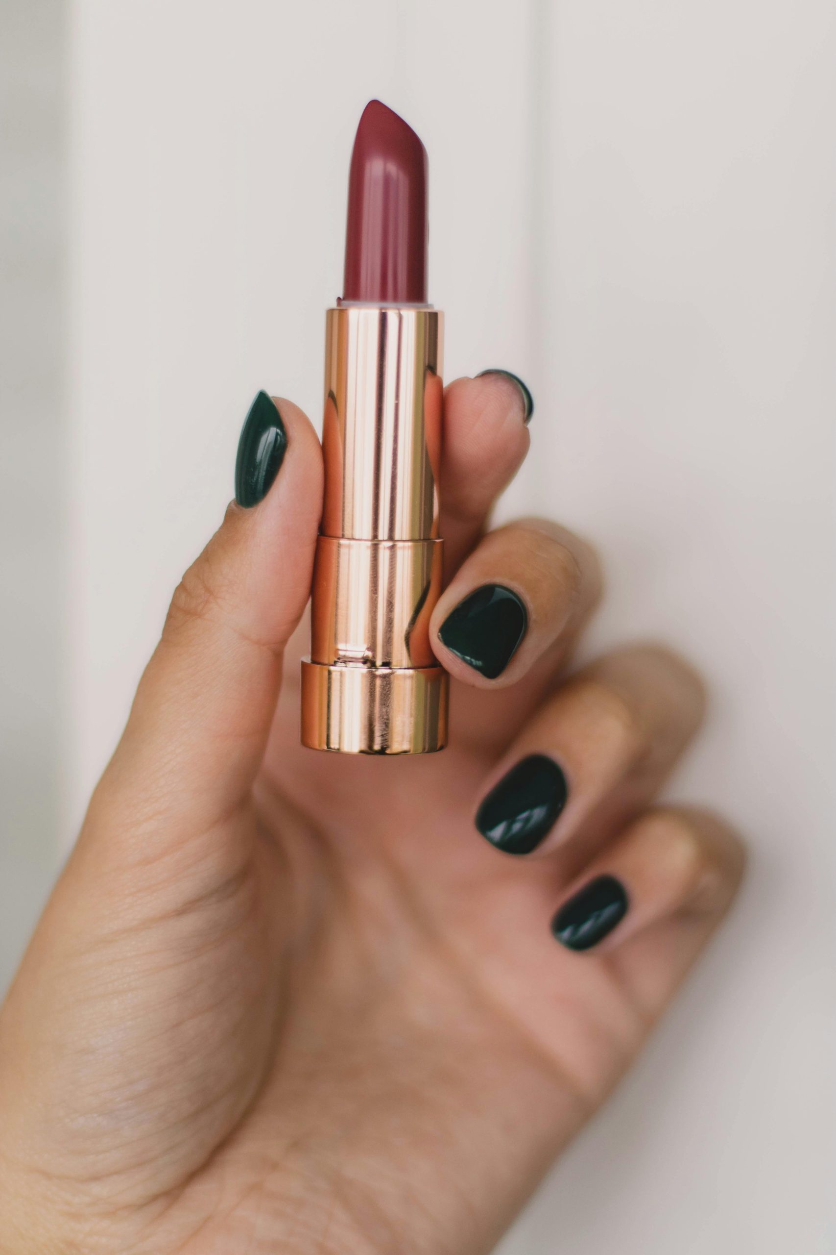 Close-up of a hand with dark polished nails holding a stylish lipstick tube against a neutral background.